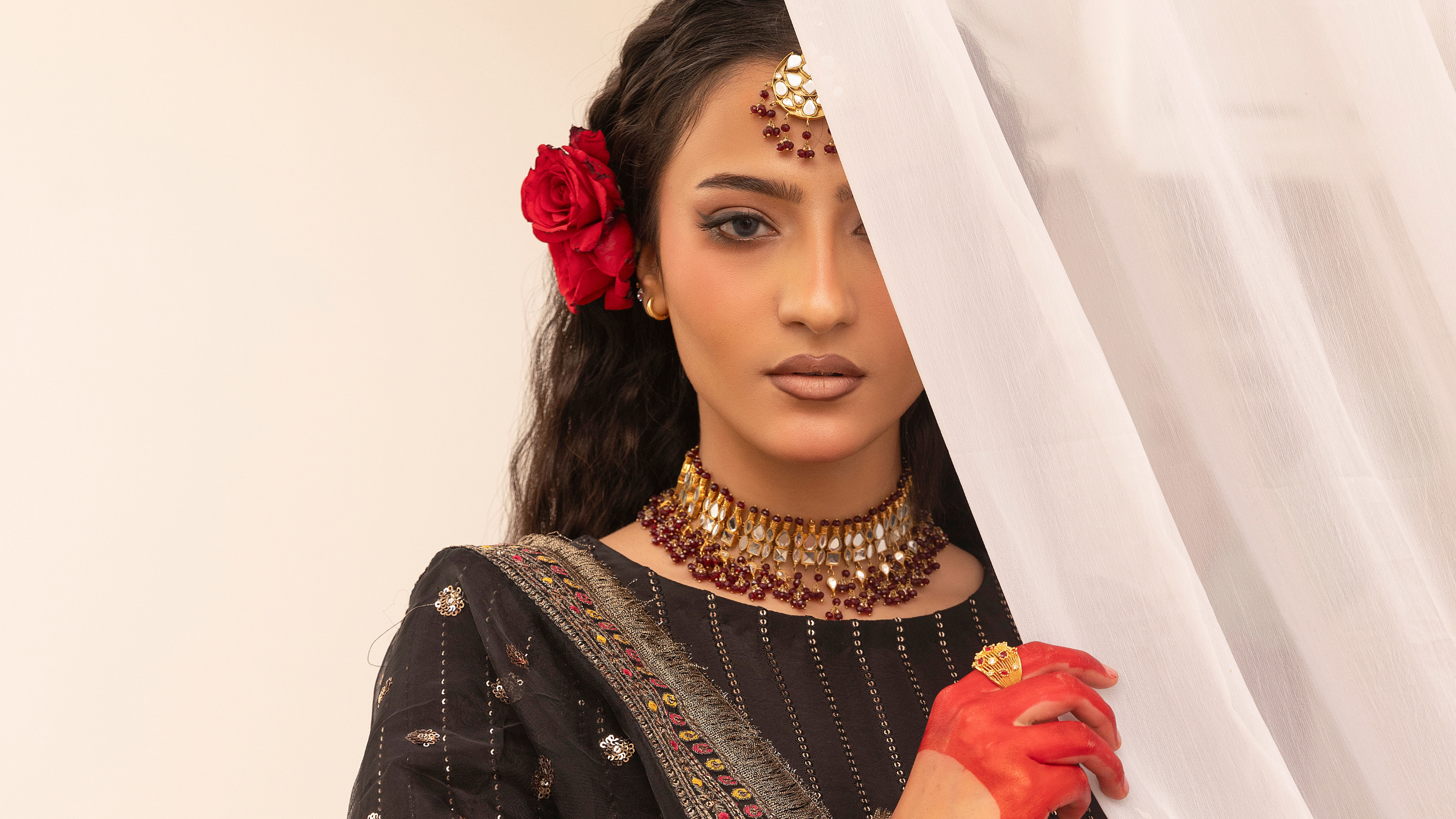 Woman in traditional attire with jewelry and a red flower in her hair, holding a white curtain.